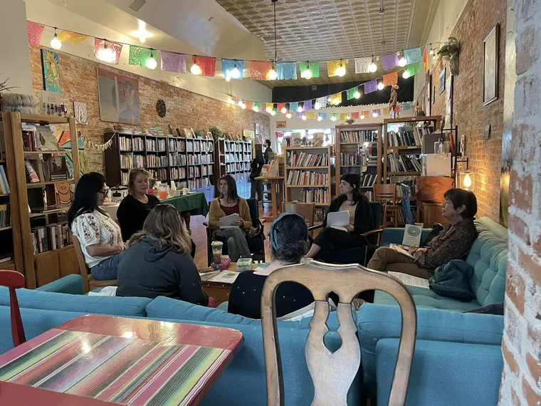 The club in session: seven readers in a circle of mismatched chairs and sofas in the back room, pages in hand, festoon lights on