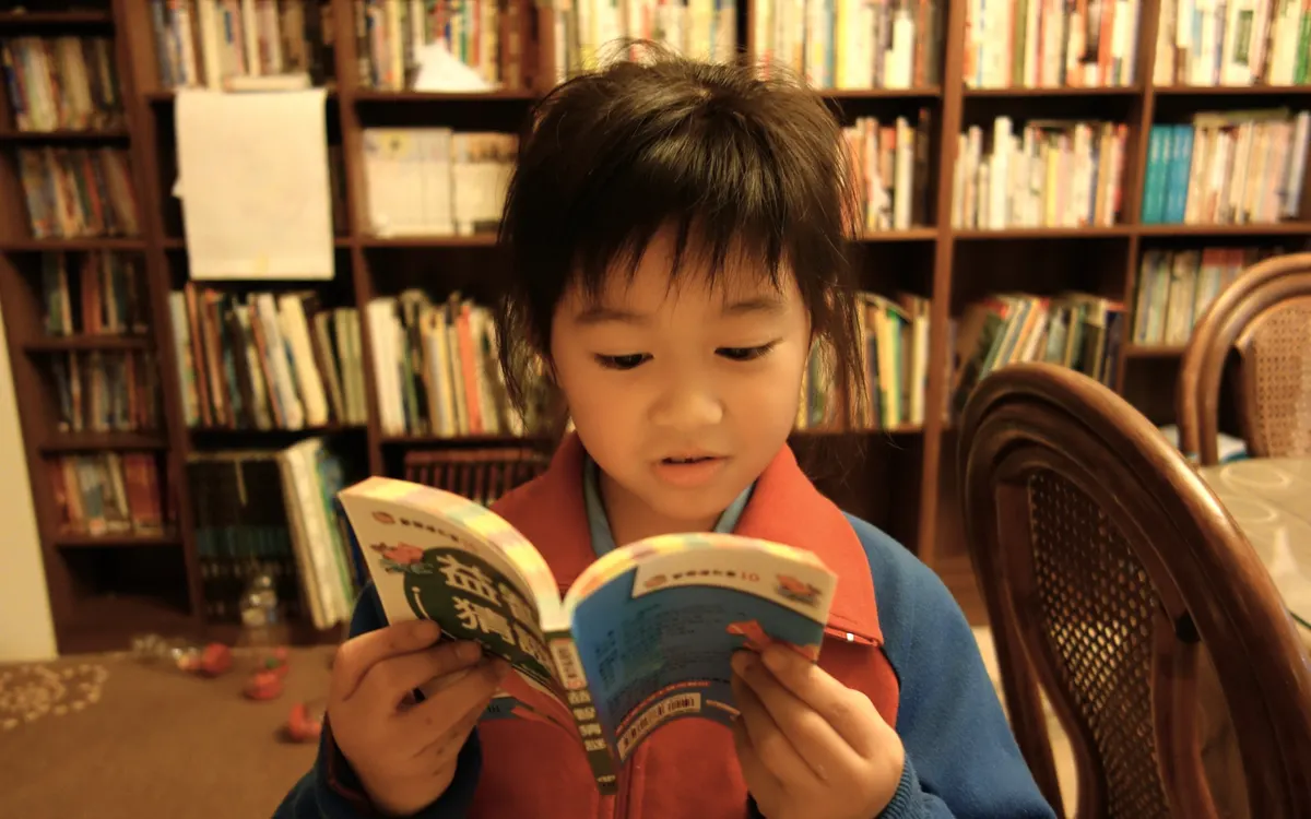 A young reader at storytime, book open in both hands, mid-sentence, the shelves full behind them