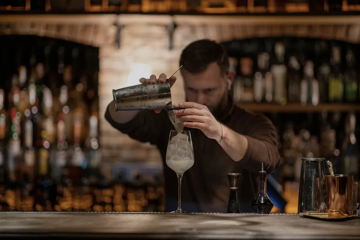 A bartender straining a stirred cocktail into a coupe behind the bar, warm lamplight and shelves of bottles in the background.