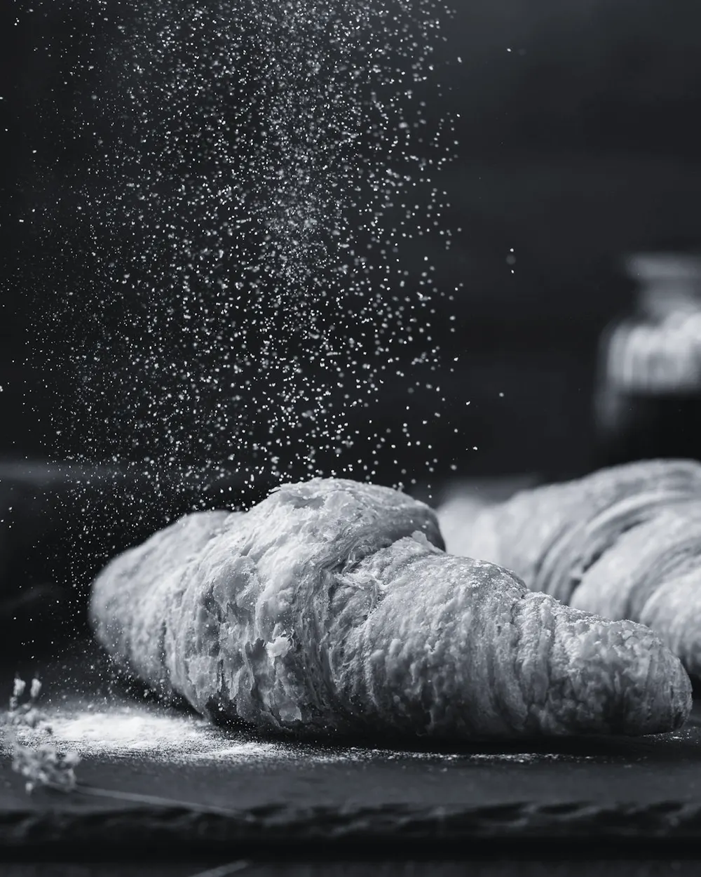 Croissants on a bakery bench under falling flour, photographed in near darkness