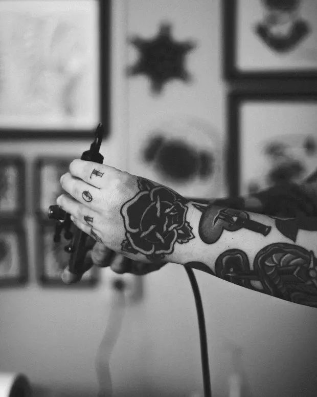 Fen Adler's tattooed hands holding a liner machine in front of framed flash