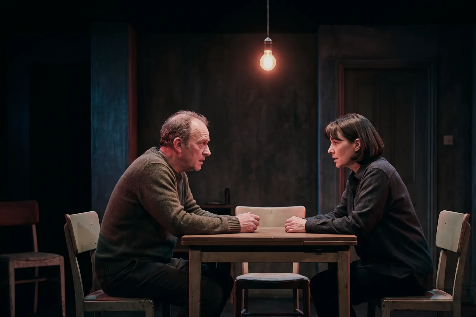 Two performers seated across a modest dining table under a single warm stage light.